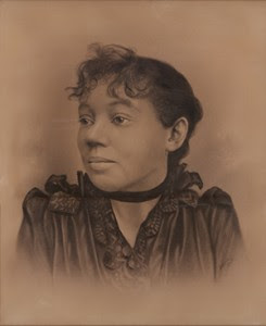 Bust image of Elizabeth Carter Brooks - woman with dark hair in an up-do, wearing a dark top with a dark ribbon around her neck and shrugging her shoulders.