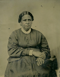 Photograph of Amelia Piper - woman with dark hair in an up-do. Woman wearing a dark-colored skirt and top with white collar and shirt cuffs. Woman sitting slightly hunched with both hands on her left waist/thigh.