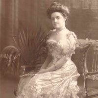 Photograph Of Cara Leland Rogers - Young Woman In Elaborate Embroidered Light-colored Dress. Woman Is Sitting On An Elegant Bench And Has Hair In An Updo.