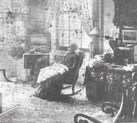 Photograph of Cornelia Grinnell - older woman in a dark dress sitting in a rocking chair inside a home, possibly a living room.