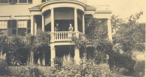 A large Greek revival style house with many plants growing around it. On a covered porch, there are two women standing.