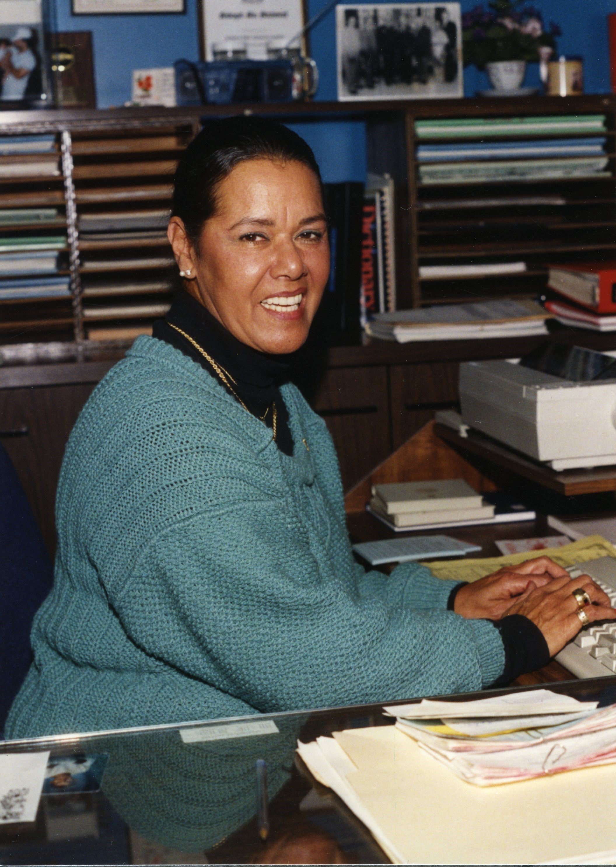 Photograph Of Marial Harper - A Woman With Black Pulled Back Hair, She Is Wearing A Blue Sweater And Typing On A Computer Keyboard