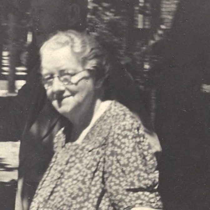Photograph of Mary Ann Hayden - an older woman with short grey hair and round glasses, she is wearing a floral print dress