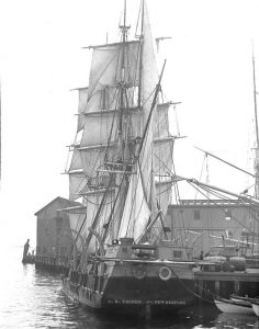 A whaling vessel tied up alongside a wharf. It is viewed from the stern and all of the sails are up. 