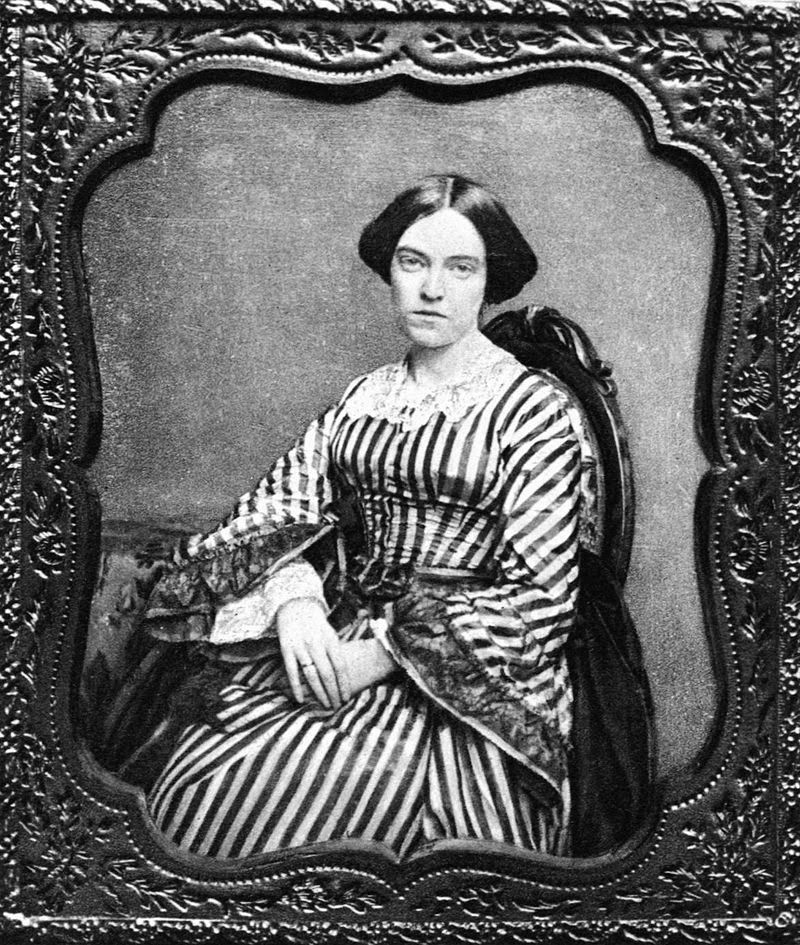 Photograph of Elizabeth Barstow Stoddard -- black and white of woman sitting in a chair. She has her dark hair in an updo and is wearing a white and dark-color vertically-striped dress. The dress has white lace around the collar and white and dark-color lace around the cuffs.