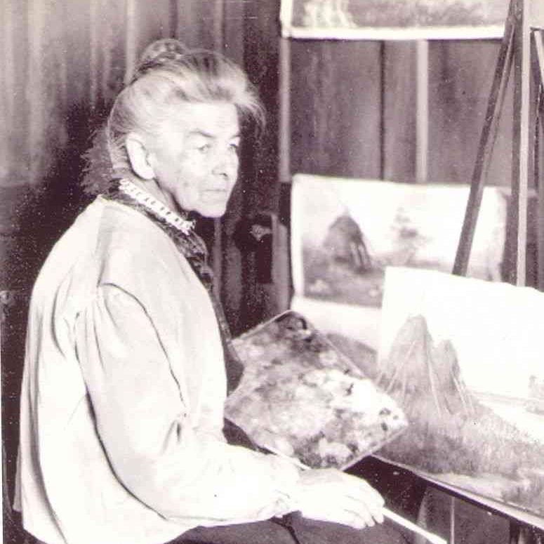 Photograph of Elizabeth Terry Delano - woman with her righ side to the camera. She sits in front of an easel with a painting of a mountain on it. She has a paint pallet and paintbrush in her hands. She has her light color hair in an up-do and she's wearing a dark dress with a white collar and a light-color jacket.
