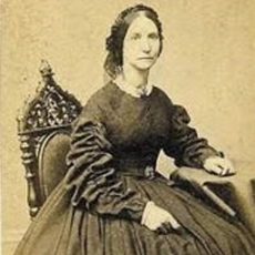 Photograph Of Ellen Kempton - A Woman With Dark Hair Pulled Back And A Dark Dress. She Has One Arm Resting On A Table.