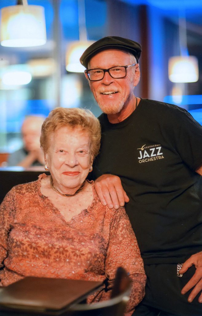 Photograph of Mary Schwartz - an older woman with short red hair and a floral shirt, smiling at the camera. She is accompanied by and older gentleman, wearing a black cap and 'Soutchcoast Jazz Orchestra' t-shirt