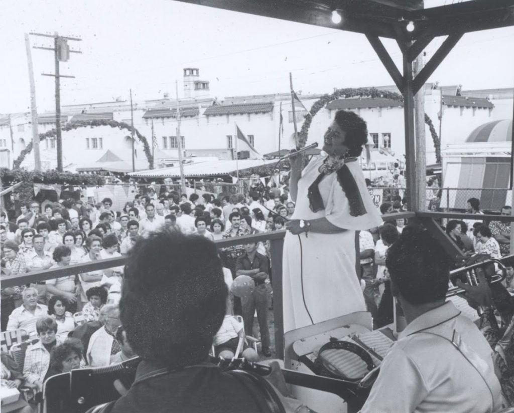 Black and white photograph of Maria Alves - a woman with black hair in a white dress -- singing on a stage with a band. In the background are many people listening to her.