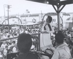 A woman with black hair in a white dress singing. In the background are many people listening to her. 