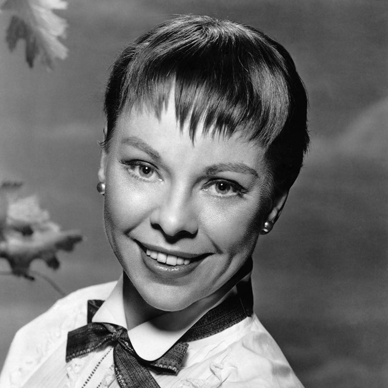 Photograph of Carol Haney - young woman smiling at the camera wearing a ribbon around her collar and dark-colored pearl earrings. Woman has pixie cut hair with short uneven bangs.