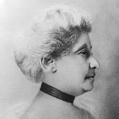 Photograph Of Elizabeth Piper Ensley - Profile Of Woman. She Has Light Color Hair In An Up-do And She's Wearing A Black Choker Necklace And Some Kind Of See-through White Wrap Around Her Shoulders.