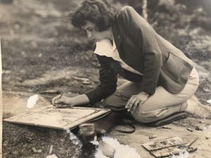 A white woman with short brown hair kneeling. She is leaning over and drawing on a pad of paper. 