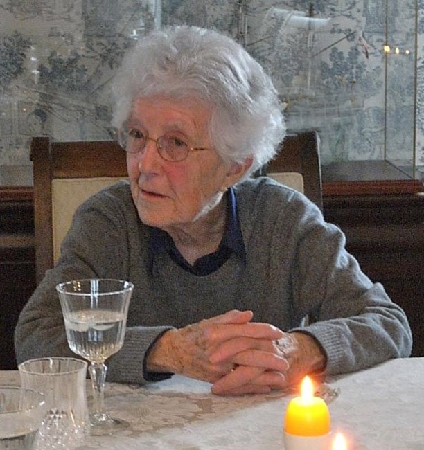 Photograph Of Hope Atkinson - An Older Woman With Short Grey Hair, Round Glasses, And A Grey Sweater. She Is Sitting At A Table With Folded Hands