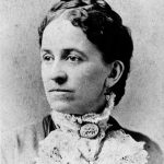 Photograph Of Bust Of Woman In Brown Towns. Woman Is Wearing Earrings And A Lace Collar Tied With A Brooch.