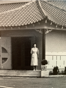 Black and white photo of a young woman in a dress standing in front of a building with a pagoda styel roof.
